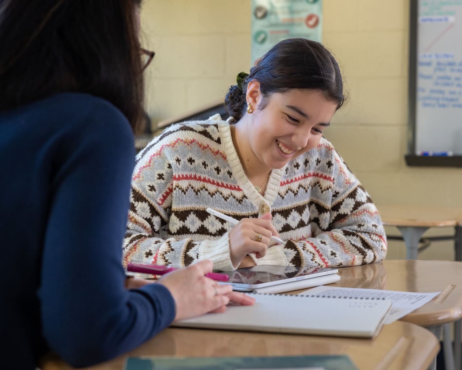 Student sitting at a desk next to a teacher while holding an Apple pencil and looking at an iPad
