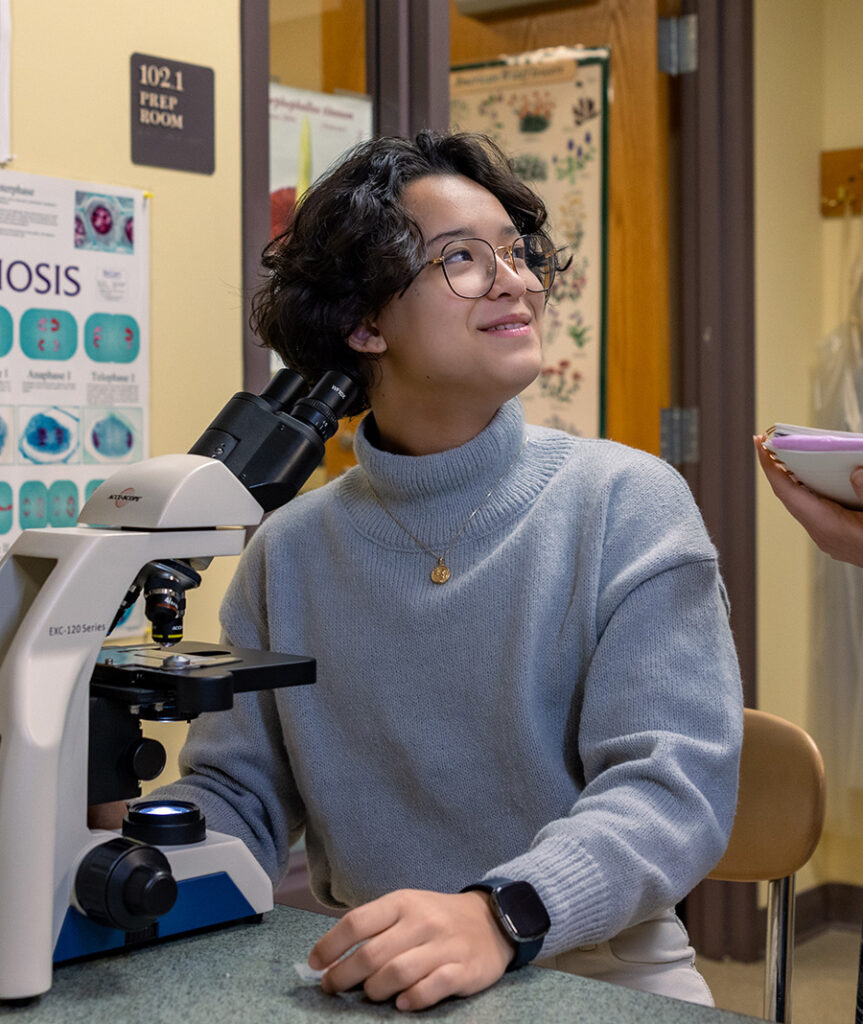 Student sitting behind a microscope while sitting at a desk and looking up at someone standing next to them