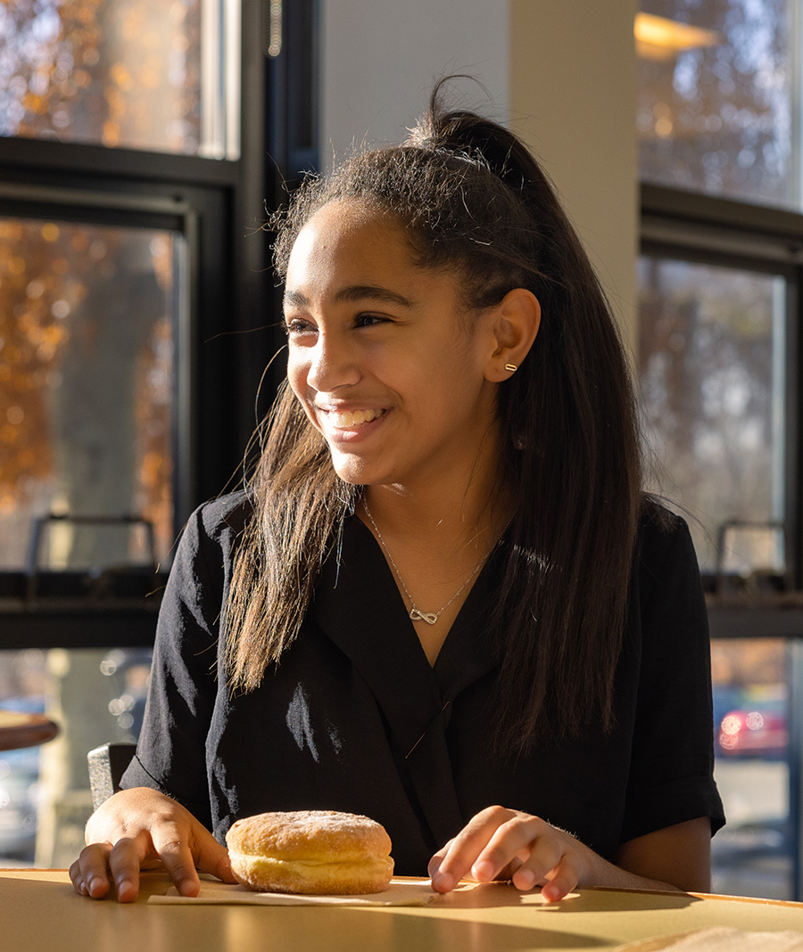 Student smiling and sitting inside in front of a window with a donut on a table in front of them
