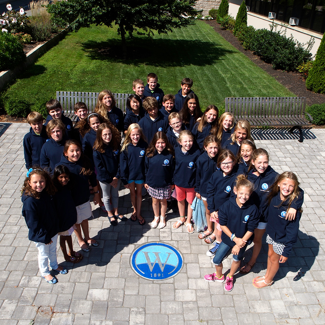Grade-school students all in matching Williams school sweatshirts and standing around Williams 1891 plaque outside.