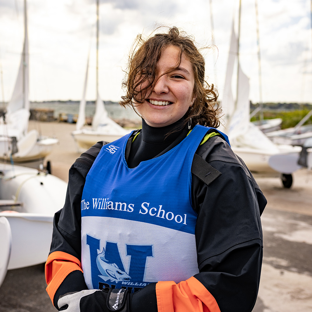 Williams School sailing student in full sailing gear smiling in front of the sailboats in the marina